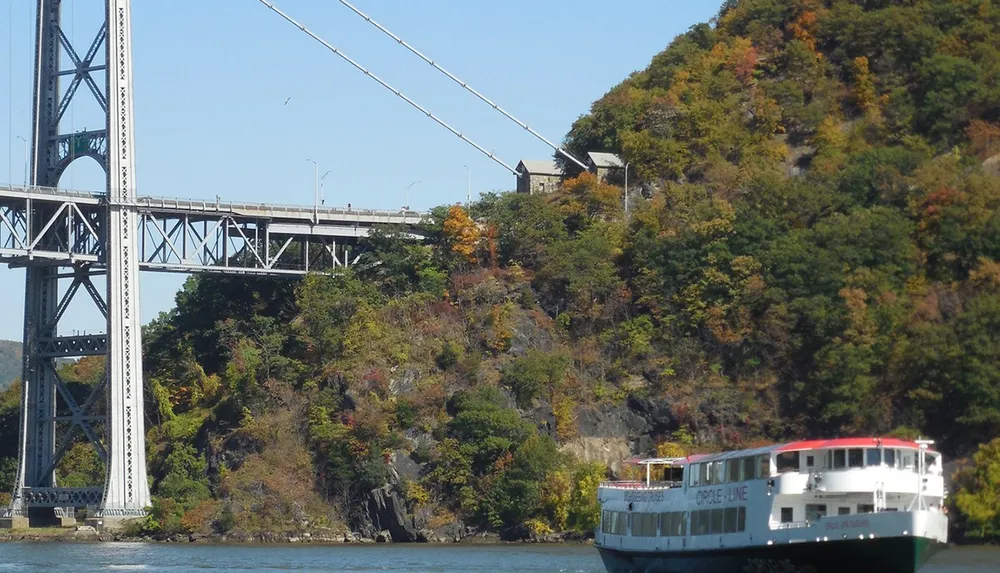 A sightseeing boat is cruising along a river near a bridge with a backdrop of a hill showing early autumn foliage