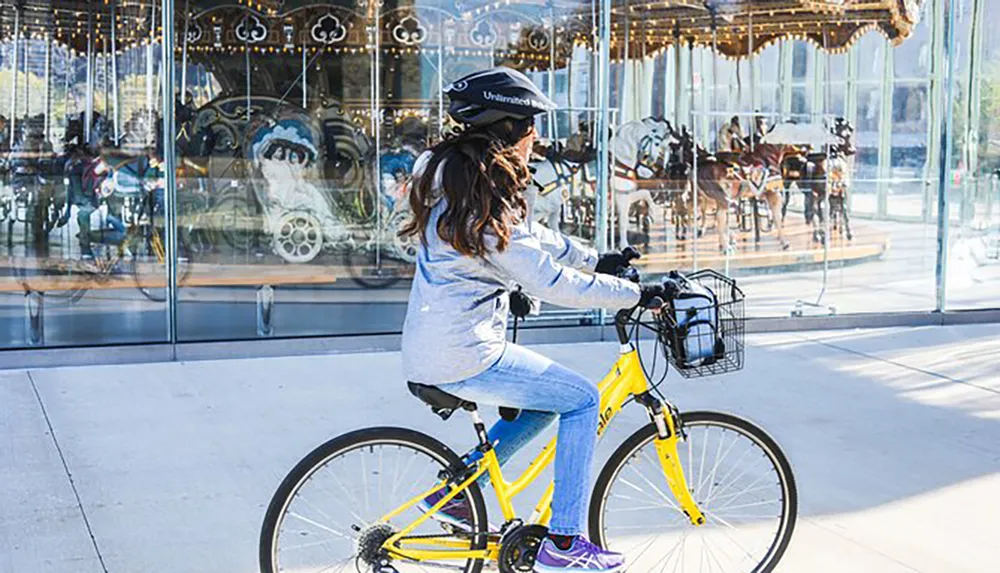 A person wearing a helmet is riding a bright yellow bicycle past a glass-enclosed carousel