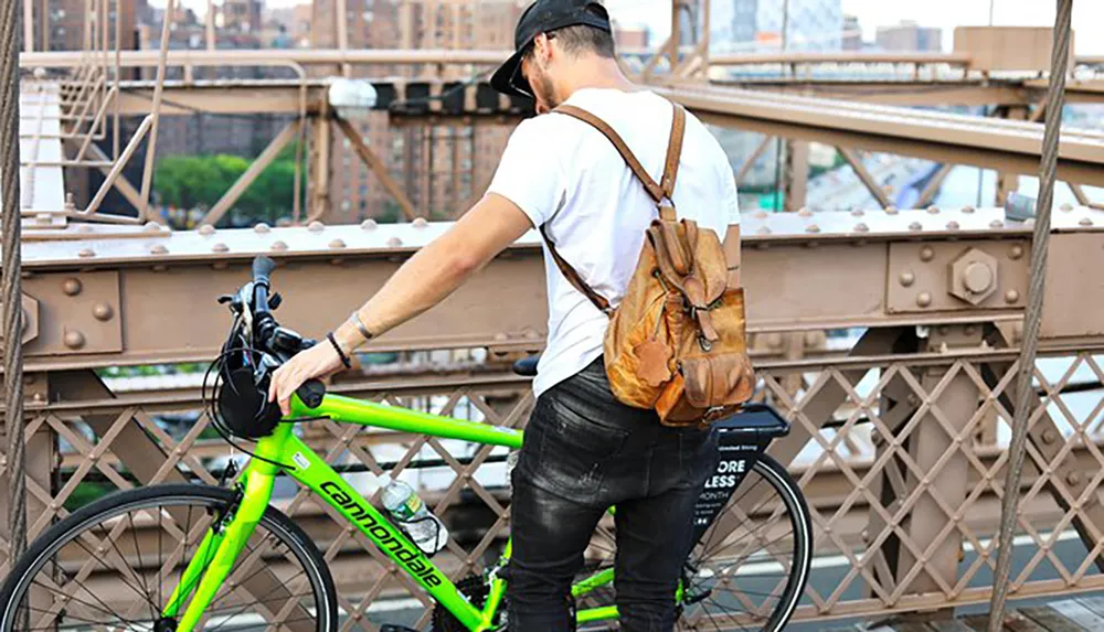 A man wearing a cap and a backpack stands beside a bright green Cannondale bicycle on a bridge with a cityscape in the background