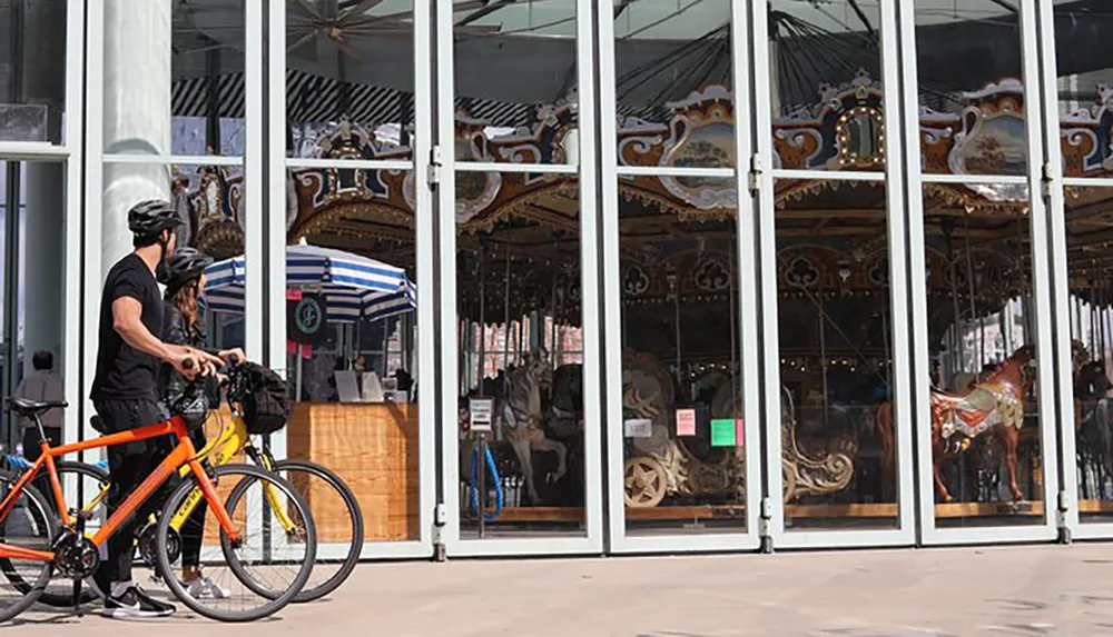 Two cyclists pause to look at a carousel enclosed within a glass facade