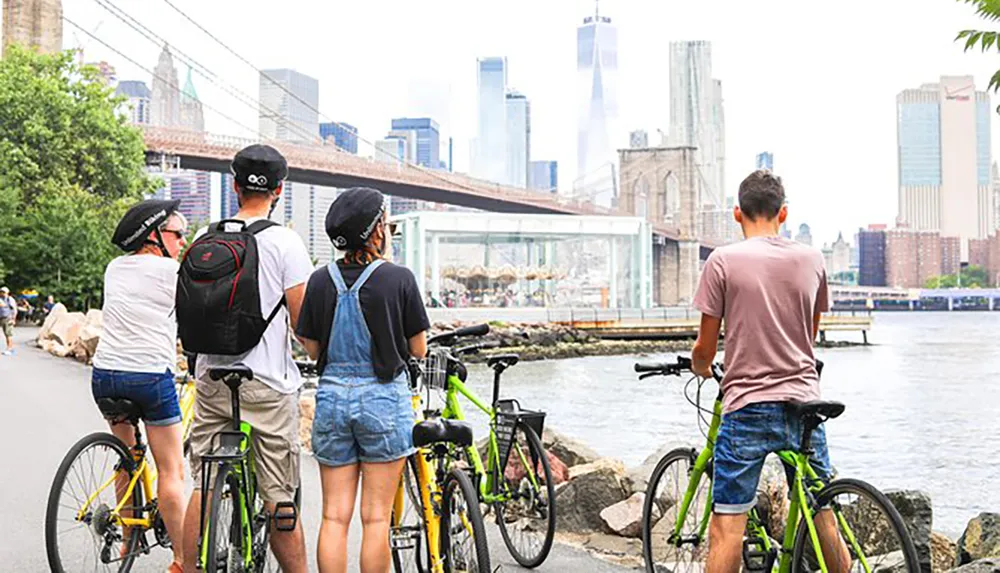 A group of cyclists is pausing to enjoy the view of the city skyline and a bridge from a riverside promenade