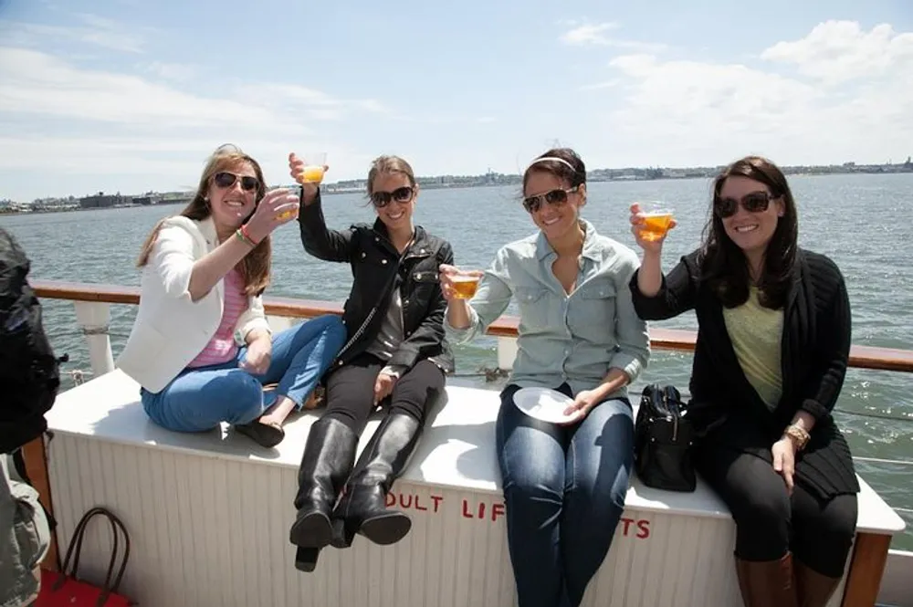 Four individuals are raising their glasses in a toast while sitting on the edge of a boat with a bright day and a body of water in the background