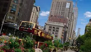 The image depicts a bustling city street scene with pedestrians, a red double-decker tour bus, and the iconic Empire State Building in the background, capturing the vibrant atmosphere of New York City.