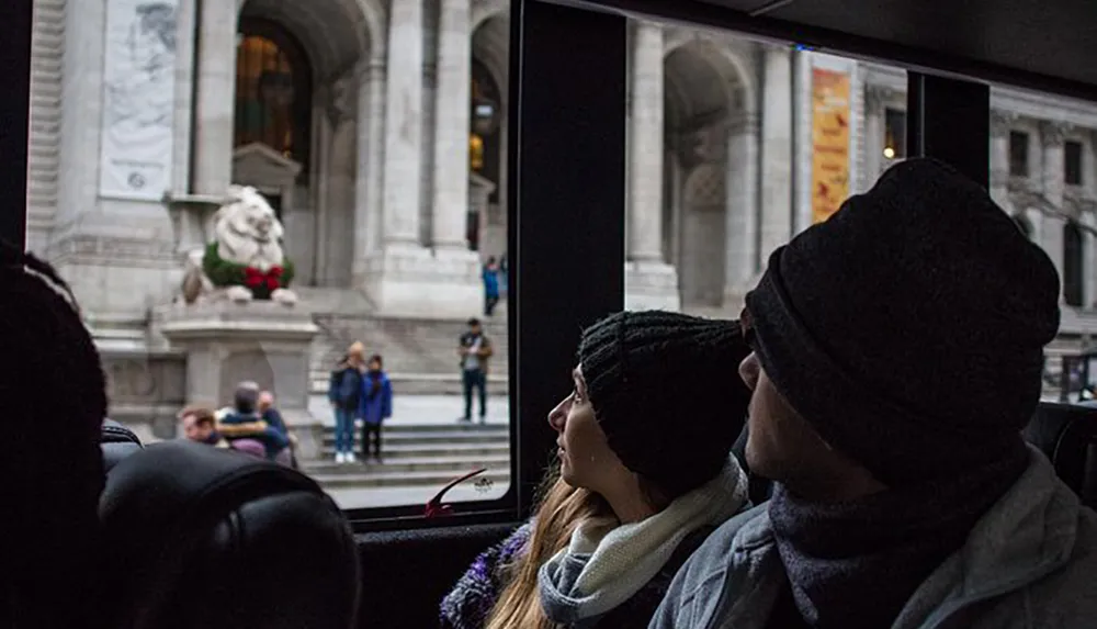 Two individuals wearing warm hats are looking out of a bus window at a building adorned with a lion statue and holiday wreaths