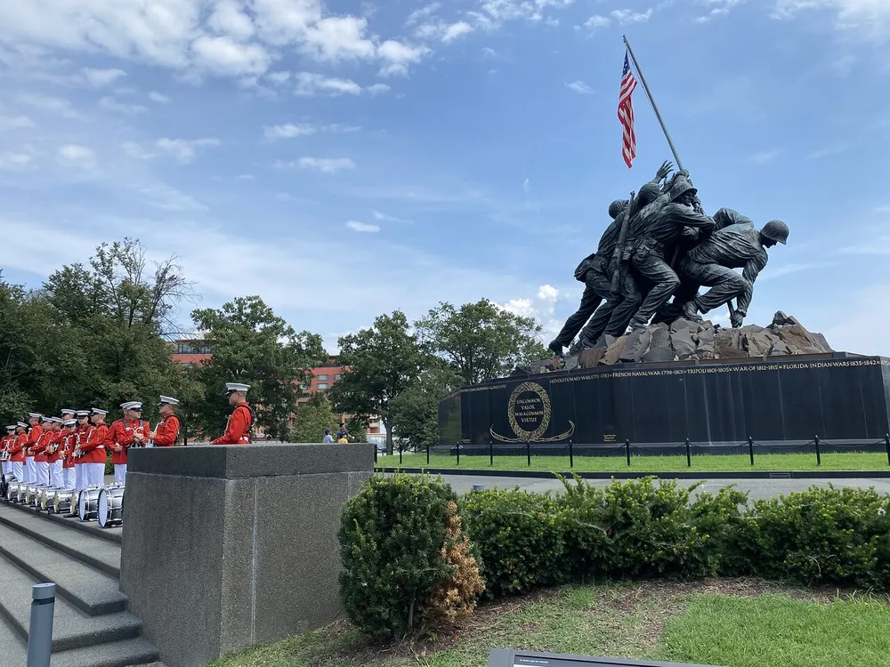 A military band in red uniforms stands in formation beside the iconic United States Marine Corps War Memorial statue against a backdrop of trees buildings and a waving American flag