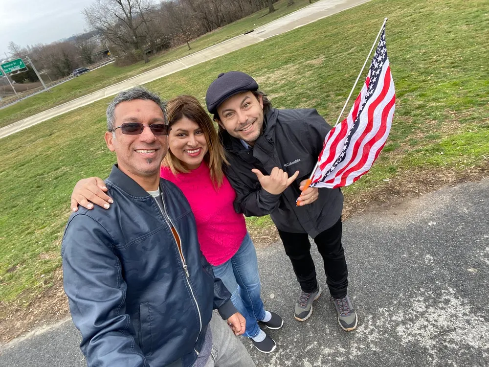 Three people are happily posing for a selfie outdoors with one of them holding an American flag