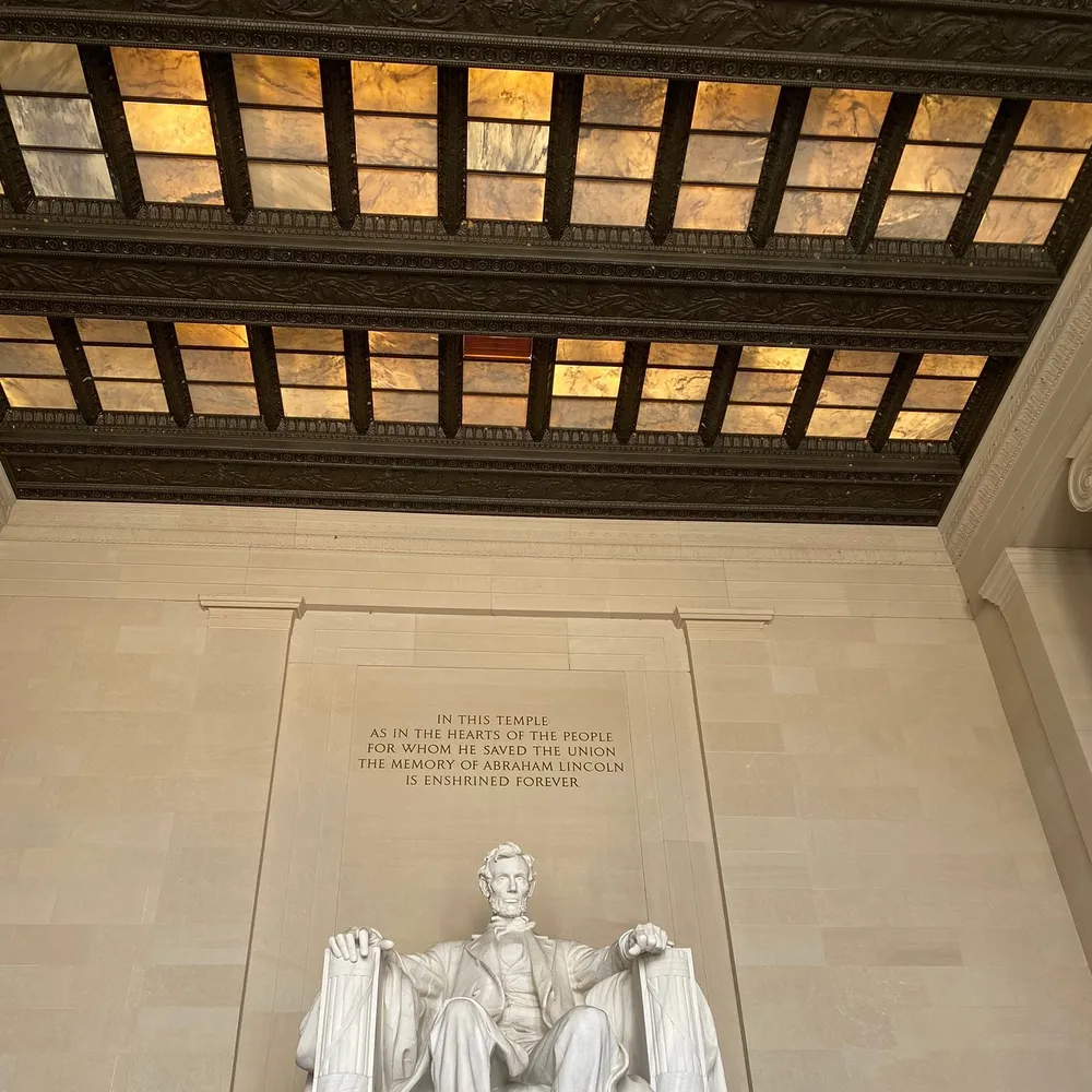 The image captures the interior of the Lincoln Memorial with the iconic statue of Abraham Lincoln and an inscription on the wall below a lit coffered ceiling
