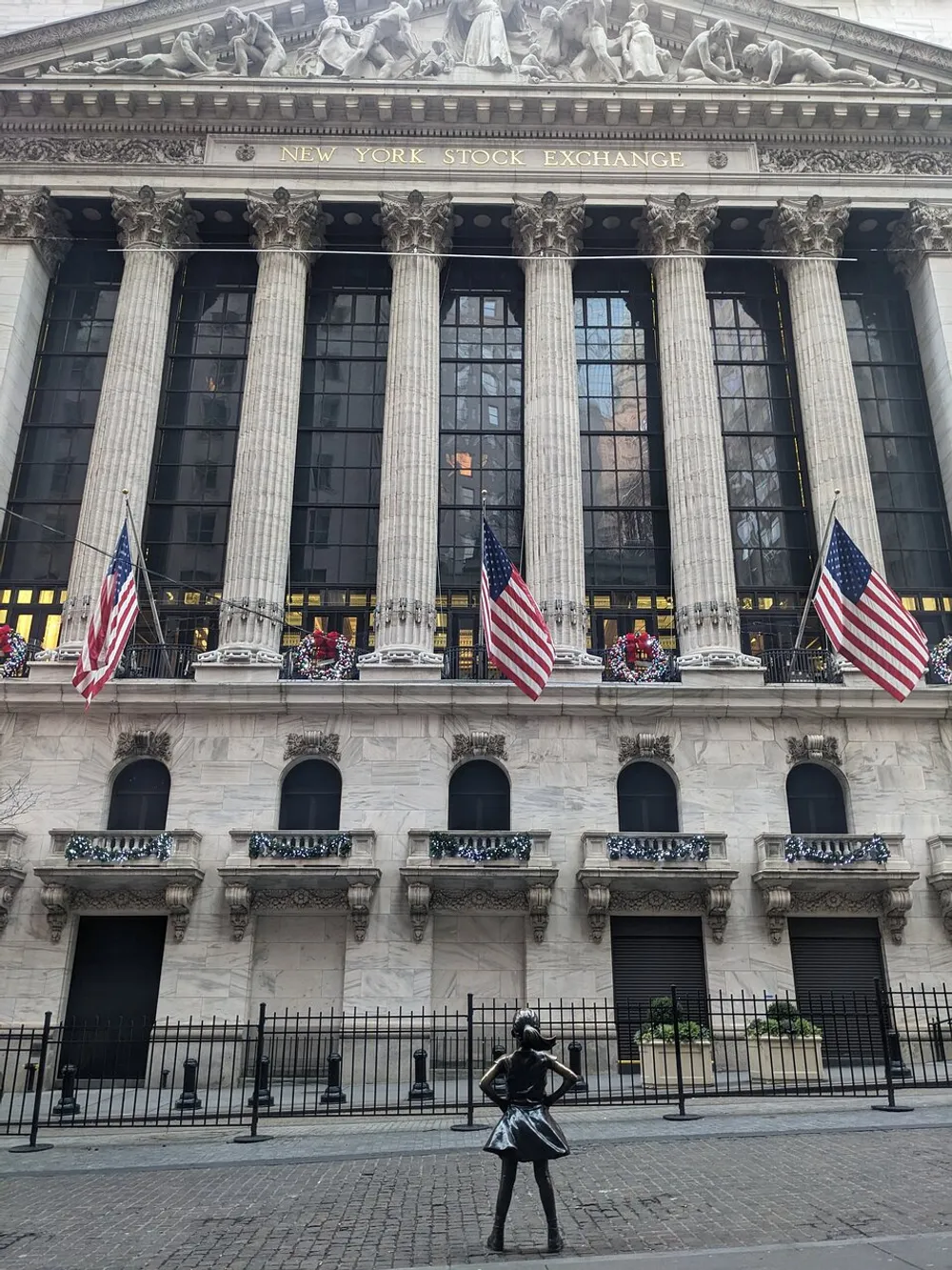 The image shows the New York Stock Exchange building with American flags and the Fearless Girl statue facing it symbolizing defiance and empowerment in the financial district