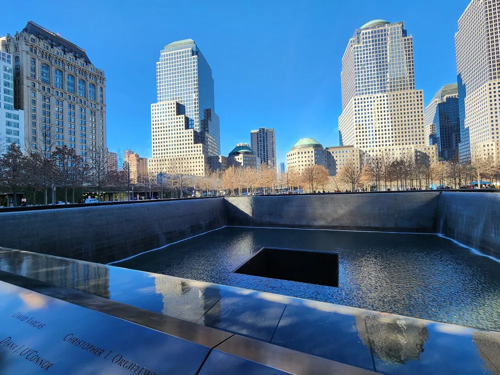 The image shows a reflective memorial pool surrounded by skyscrapers under a clear blue sky