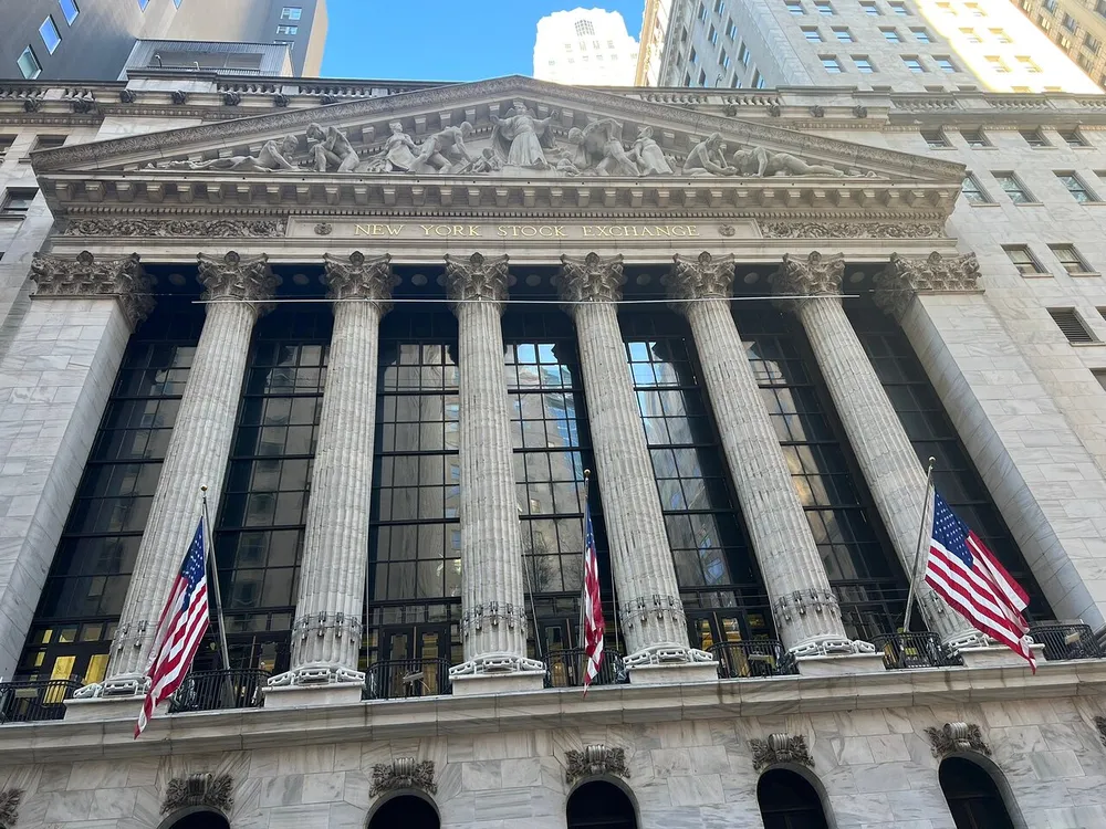 The image shows the neoclassical facade of the New York Stock Exchange adorned with American flags under a clear blue sky