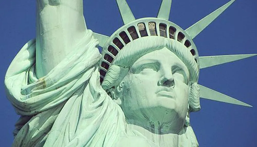 The image is a close-up of the face and crown of the Statue of Liberty against a clear blue sky