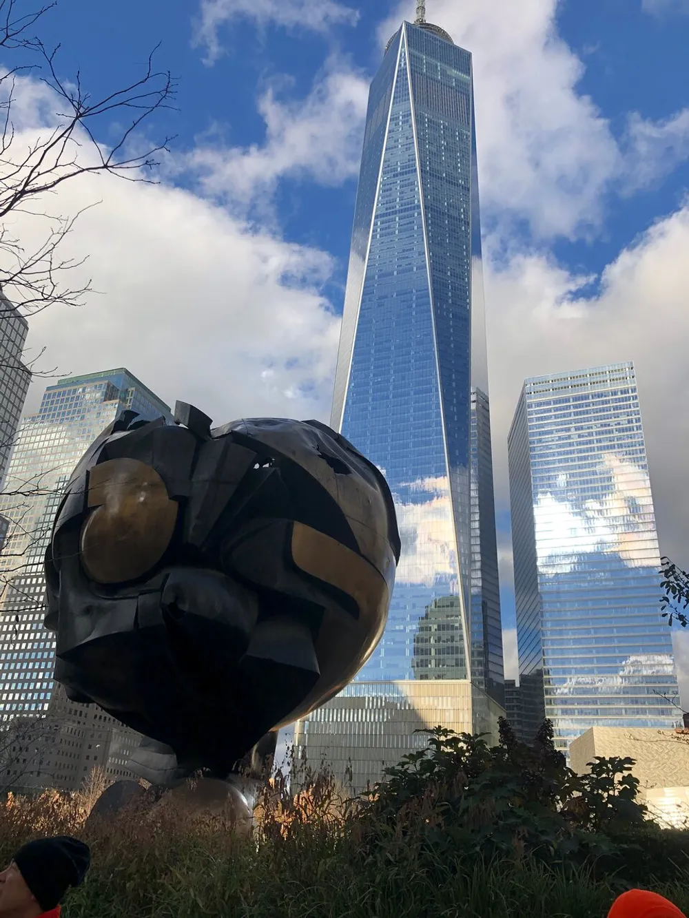 The image shows the Sphere sculpture with damage from 911 set against a backdrop of the One World Trade Center under a partly cloudy sky
