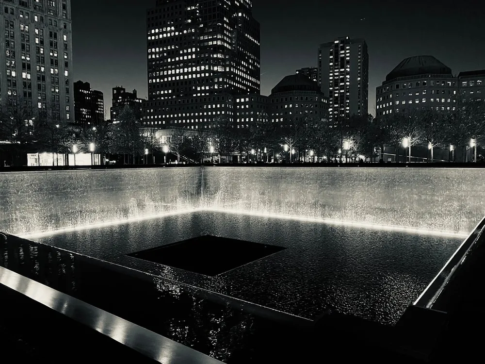 The image captures a serene black-and-white night view of the 911 Memorial in New York City featuring one of the twin reflecting pools