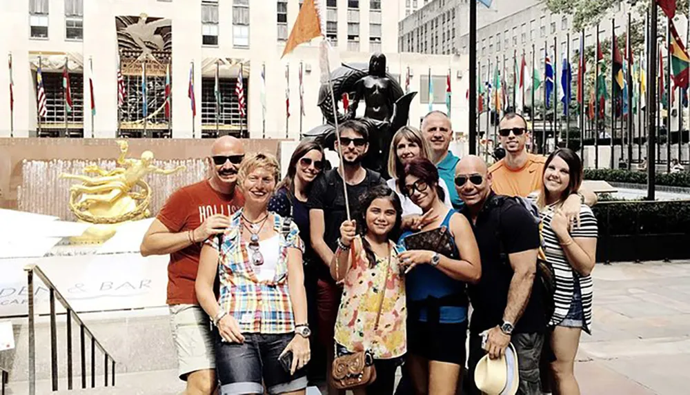 A group of people is posing for a photo in front of a dark statue of Batman at Rockefeller Center with the iconic golden Prometheus statue and international flags in the background