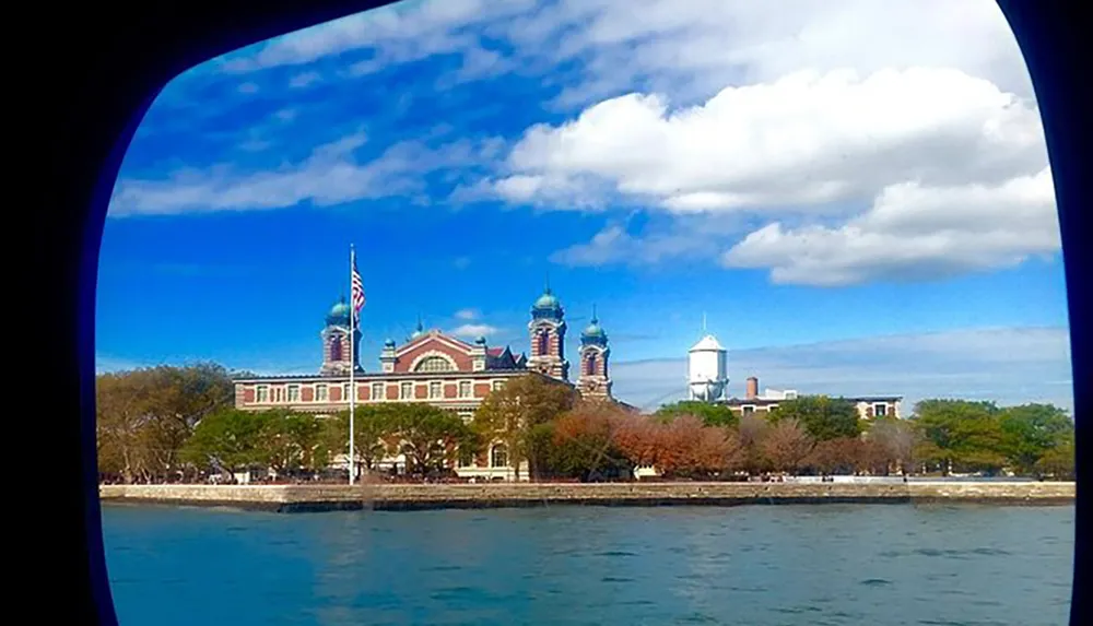 Viewed through a porthole this image captures the historic Ellis Island building under a blue sky with fluffy clouds