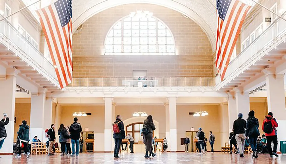 The image depicts a spacious indoor hall with high ceilings two large American flags hanging prominently and several people milling about some taking photos and others seemingly in transit