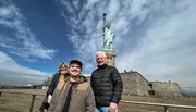 Three people are posing for a photograph with the Statue of Liberty in the background.