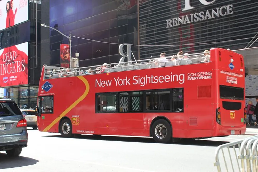 A red double-decker sightseeing tour bus with passengers on the upper deck is driving through a busy street with illuminated billboard advertisements in the background