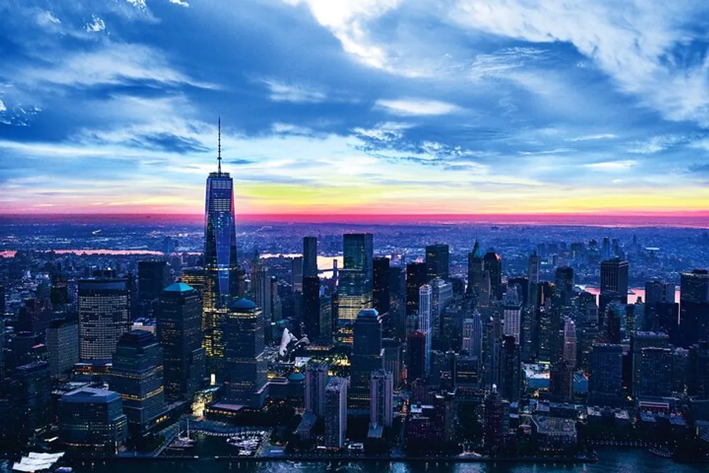 The image shows an aerial view of a densely clustered city skyline with skyscrapers punctuating the evening sky backlit by a fiery sunset