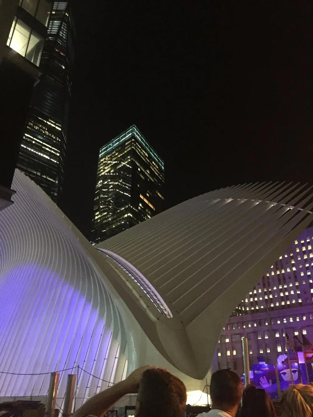 The image shows the illuminated wing-like structure of the Oculus at the World Trade Center transportation hub in New York City with skyscrapers towering in the background at night
