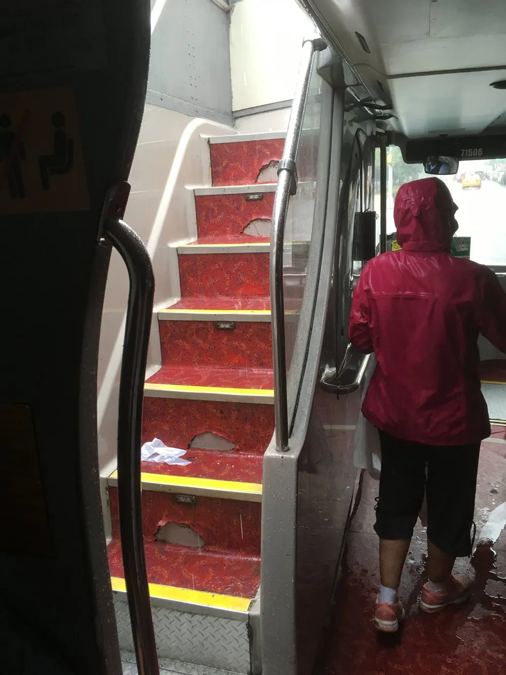 A person in a red raincoat is standing at the open door of a bus which shows a set of stairs leading to the street on a wet and possibly rainy day