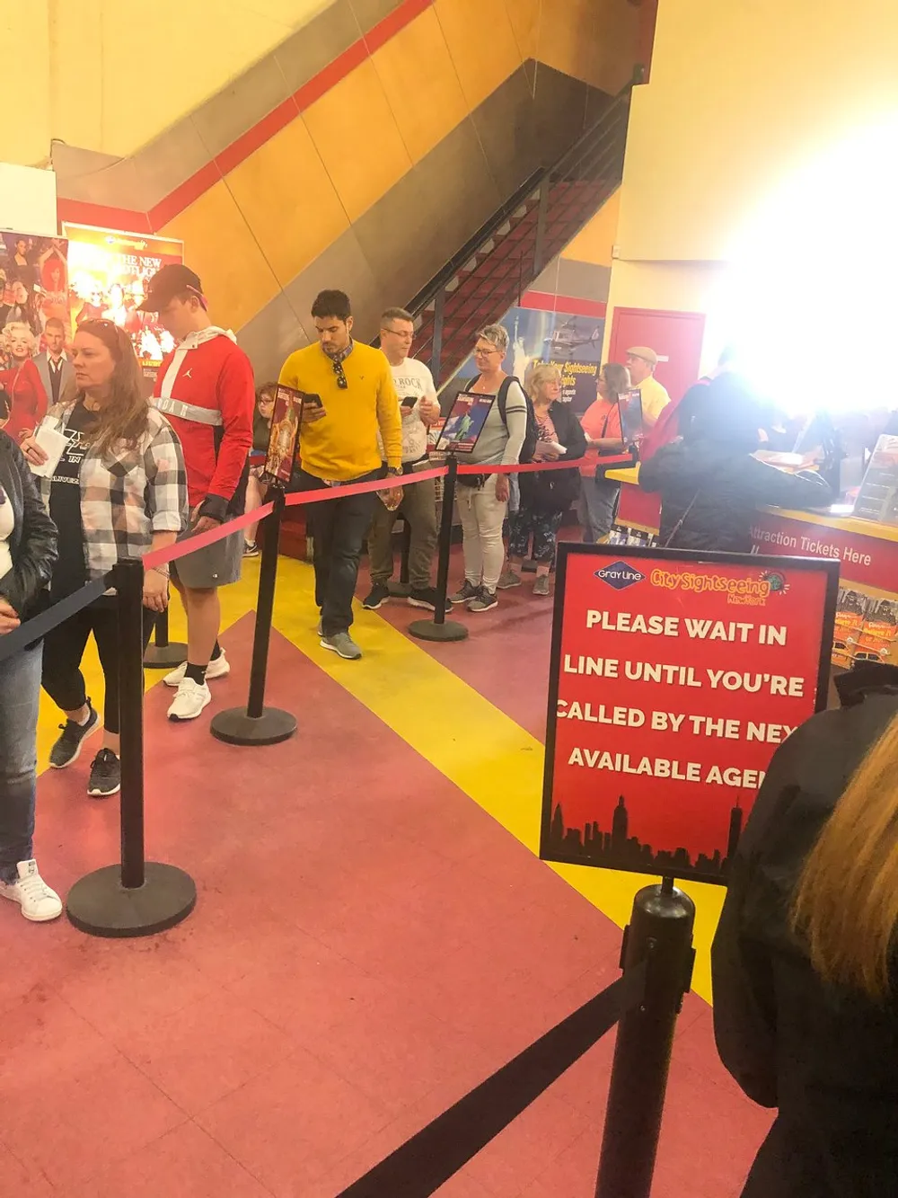 Guests are queuing indoors in front of a ticket counter with a sign indicating to wait until being called by the next available agent