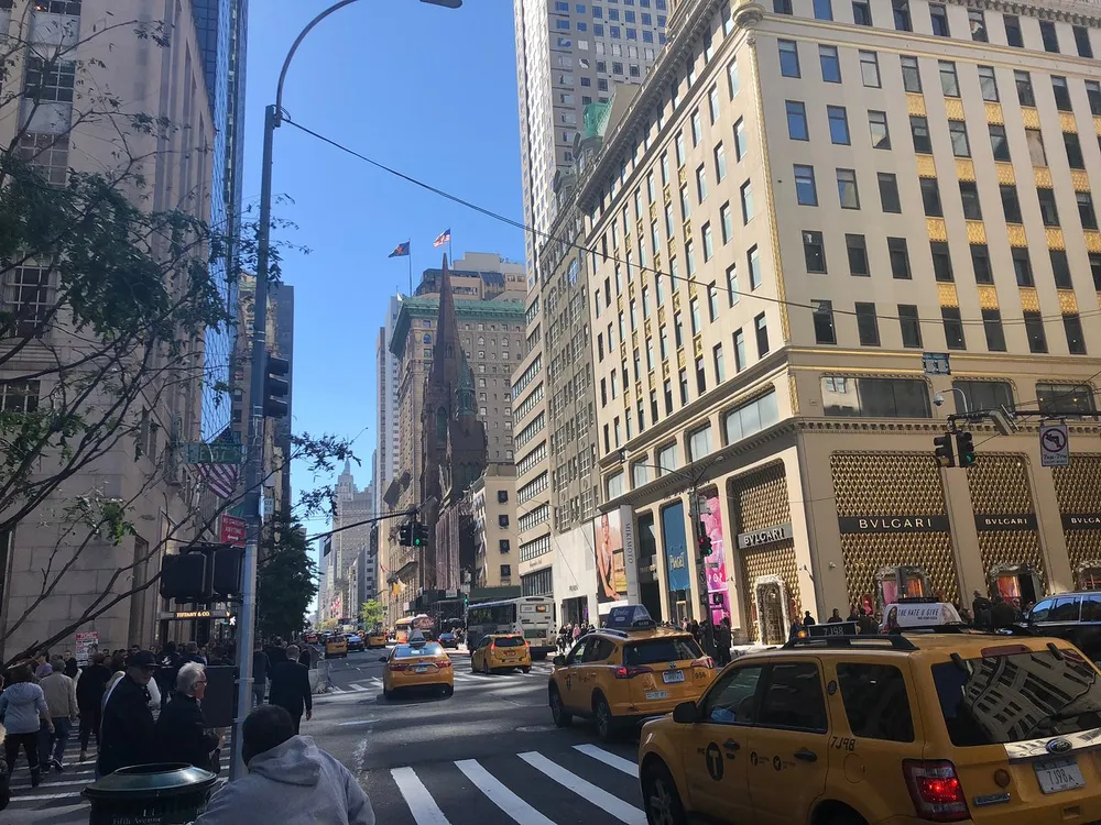 A bustling city street scene with pedestrians and yellow taxis featuring prominent commercial buildings under a clear blue sky