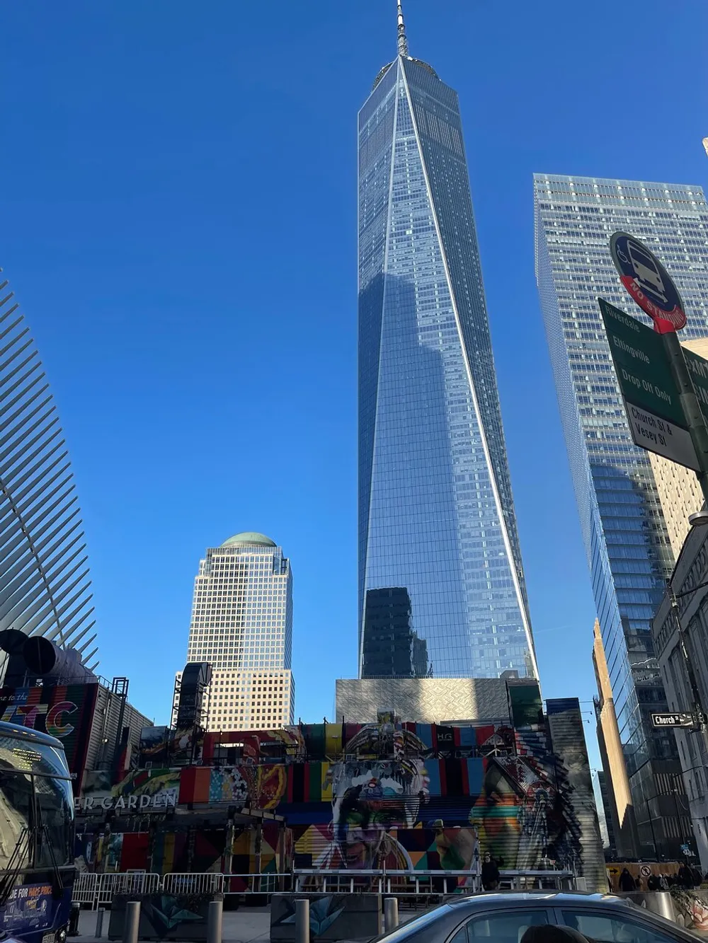 The image shows a bustling street scene with the towering One World Trade Center in the background under a clear blue sky