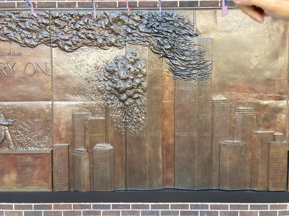 The image shows a bronze relief sculpture with a skyline textured clouds and American flags conveying a sense of commemoration or tribute