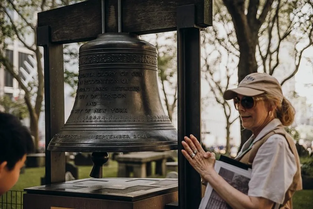 A woman wearing a cap and sunglasses admires a large bell mounted on a frame that bears an inscription with a partial view of a young person on the left