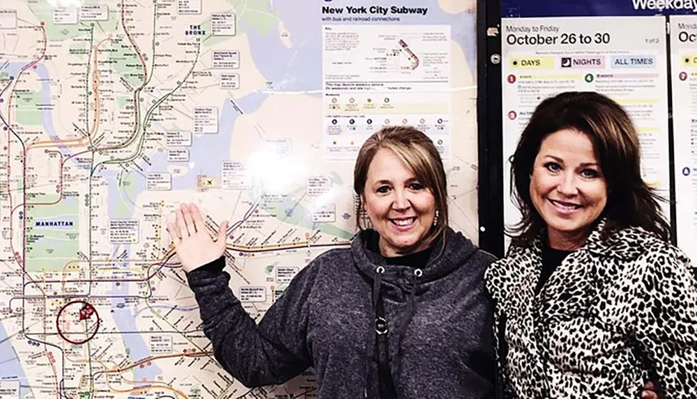 Two smiling women pose in front of a large map of the New York City subway system