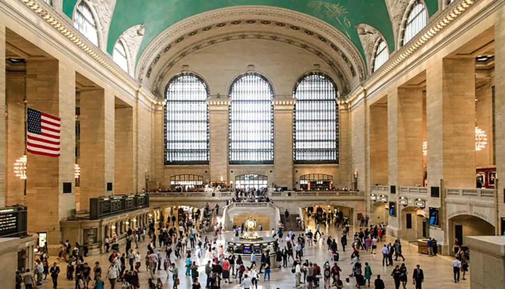 The image shows a bustling interior view of Grand Central Terminal with people commuting and an American flag displayed exemplifying the stations grand architecture and status as a transportation hub