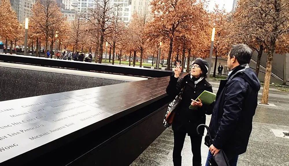 Two individuals are standing beside a reflective memorial with names inscribed on it as the woman points upwards towards the surrounding urban landscape or sky amidst a setting of bare trees and overcast weather