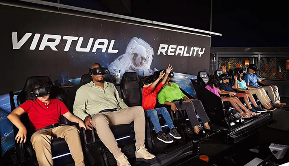A group of people is experiencing a virtual reality VR simulation in moving chairs against a backdrop that advertises VIRTUAL REALITY