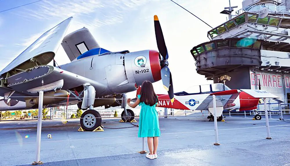 A child stands reaching out to an old military airplane on display next to a ship labeled FIGHTING I