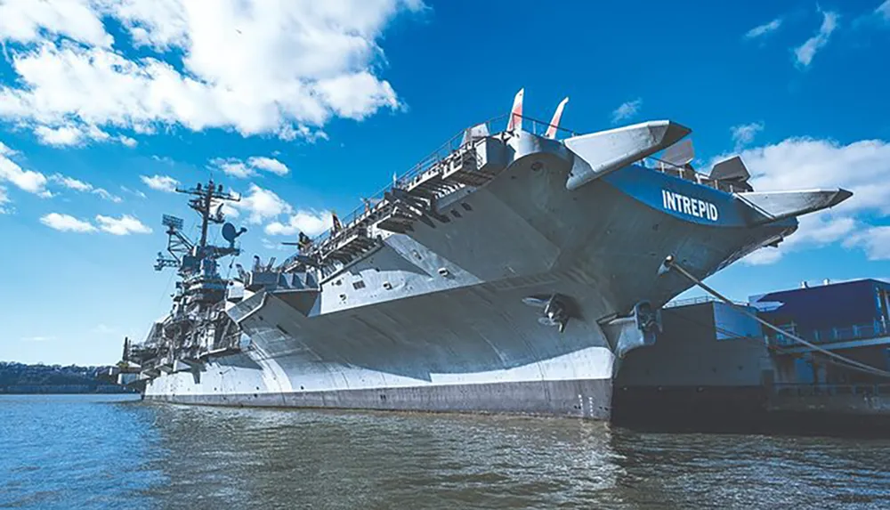 The image shows the aircraft carrier USS Intrepid now a sea air and space museum docked at a harbor under a blue sky with scattered clouds