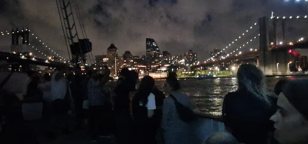 A group of people is standing by the water at night with a lit-up bridge and city skyline in the background
