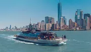 The image shows a ferry full of passengers moving across the water with the skyline of Lower Manhattan, including the One World Trade Center, in the background.