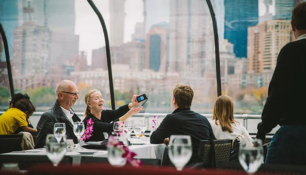 People are enjoying a meal in a riverside restaurant with a view of a cityscape with one woman taking a selfie with a man
