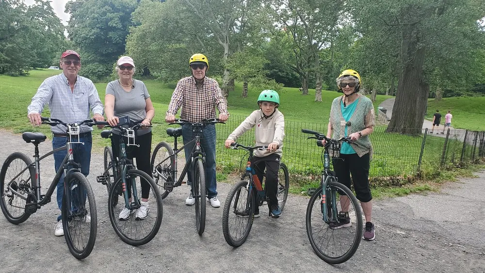 Five individuals of varying ages wearing helmets and casual attire are standing with their bicycles in a park setting preparing for a ride