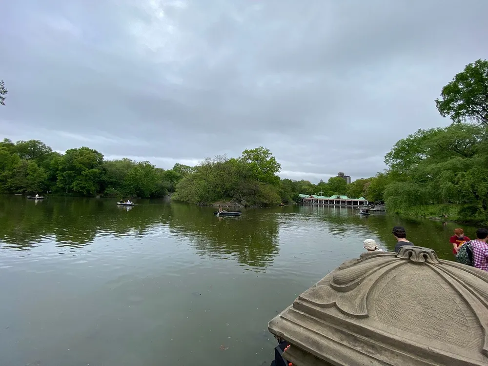 People enjoy boating on a serene lake surrounded by lush greenery under an overcast sky with the edge of a classical stone balustrade in the foreground
