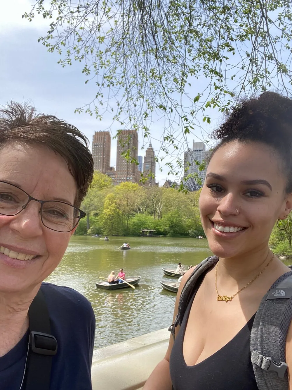 Two people are smiling for a selfie with a scenic backdrop of a lake with boaters and urban high-rises under a clear sky