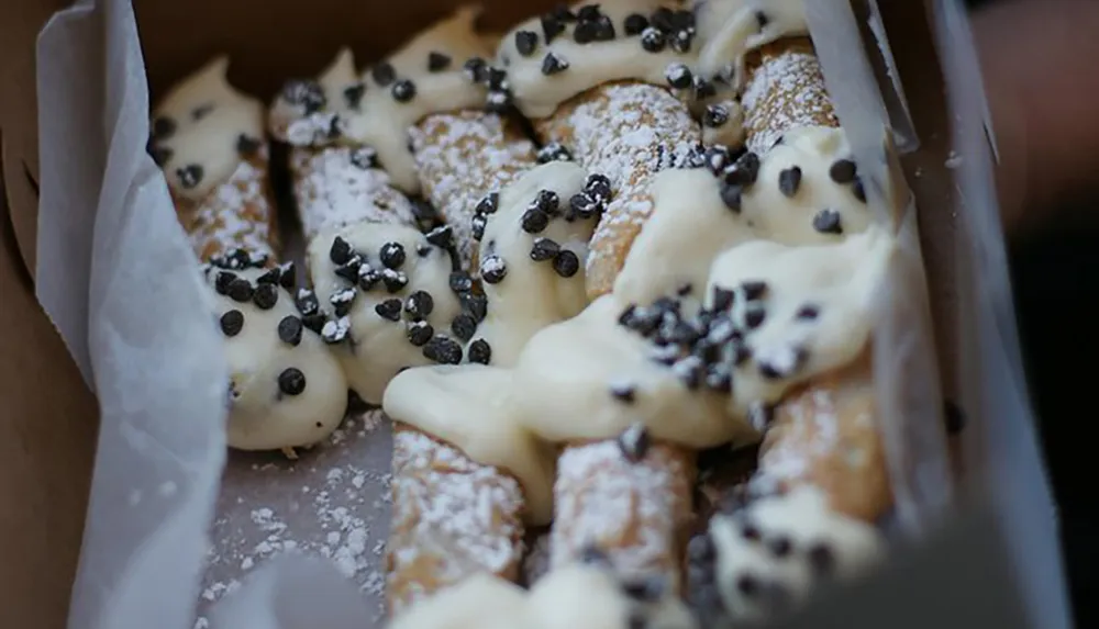 A box of freshly baked cannoli sprinkled with powdered sugar and topped with chocolate chips suggesting a delightful dessert treat