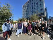 A diverse group of people poses for a photo on a sunny day with a modern building in the background.