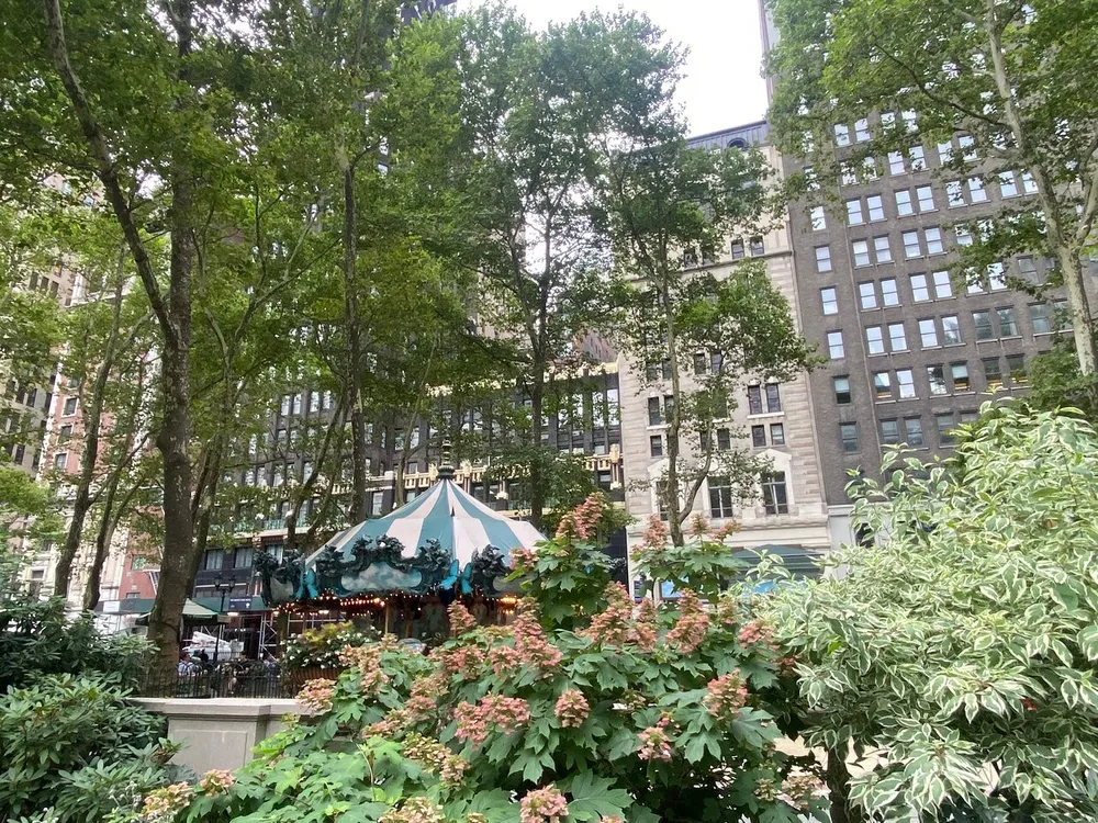The image shows a lush green urban park scene with a carousel tent amidst tall trees surrounded by high-rise buildings creating a peaceful oasis in the midst of a busy city