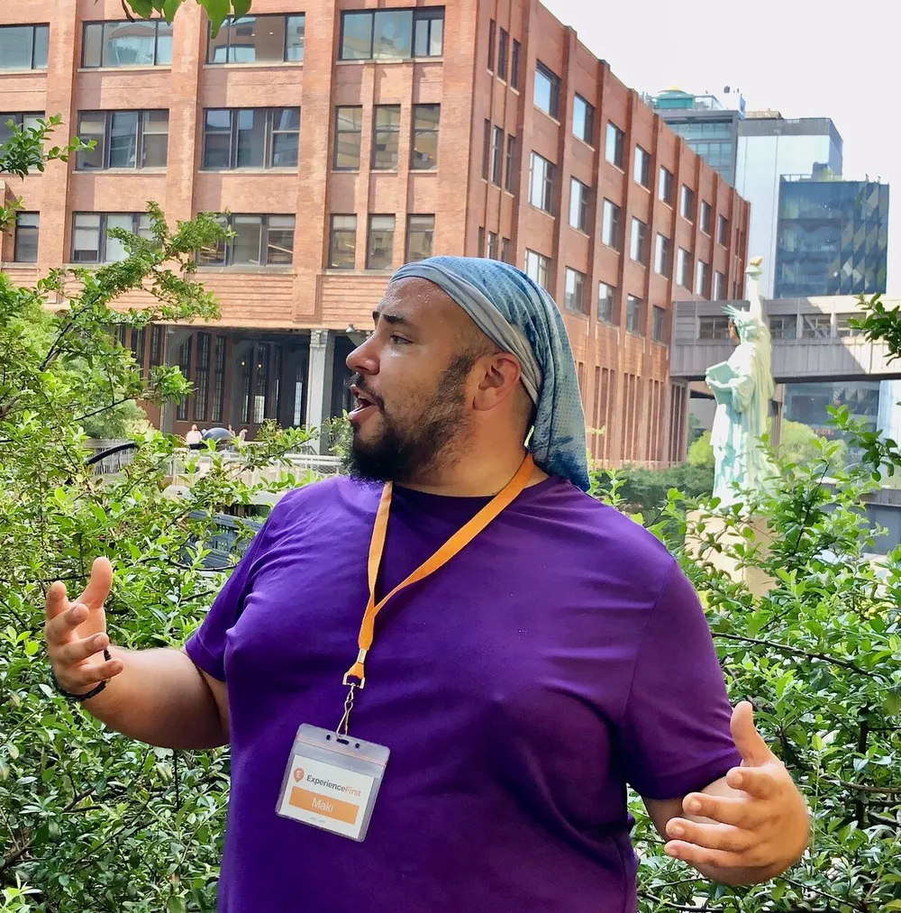 A person in a purple shirt and a headscarf is speaking and gesturing with their hands wearing a conference badge with a backdrop of buildings and greenery