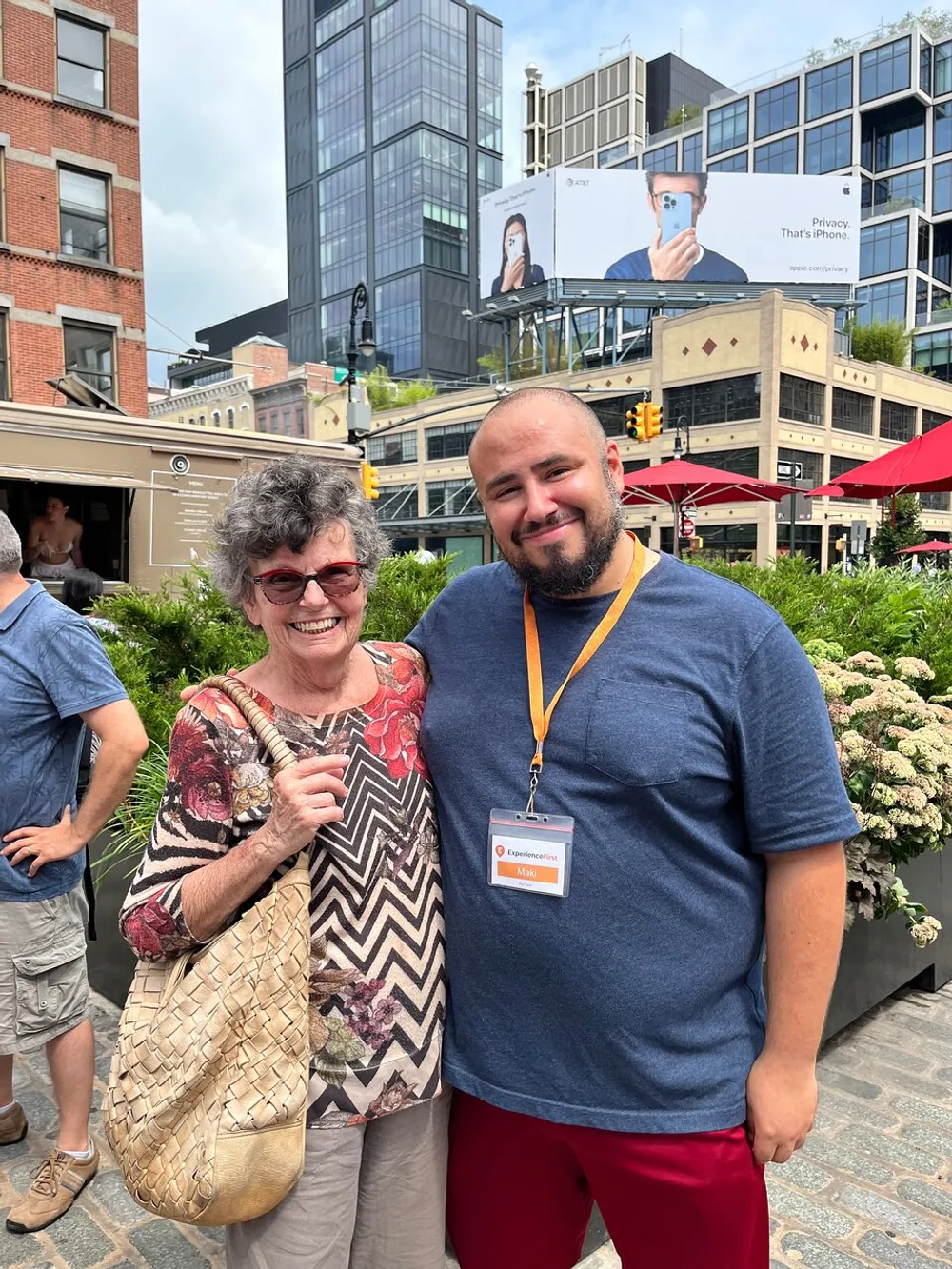 A smiling older woman and a bearded man are posing for a photo on a sunny day in an urban environment with a large iPhone advertisement in the background