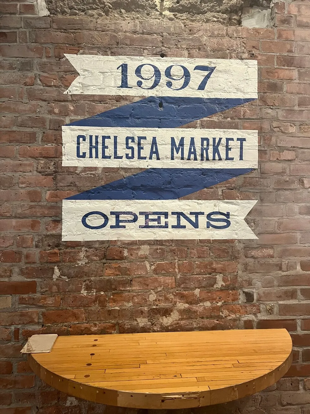 The image shows a brick wall with a painted sign that reads 1997 Chelsea Market Opens above a wooden bench indicating the opening year of the Chelsea Market