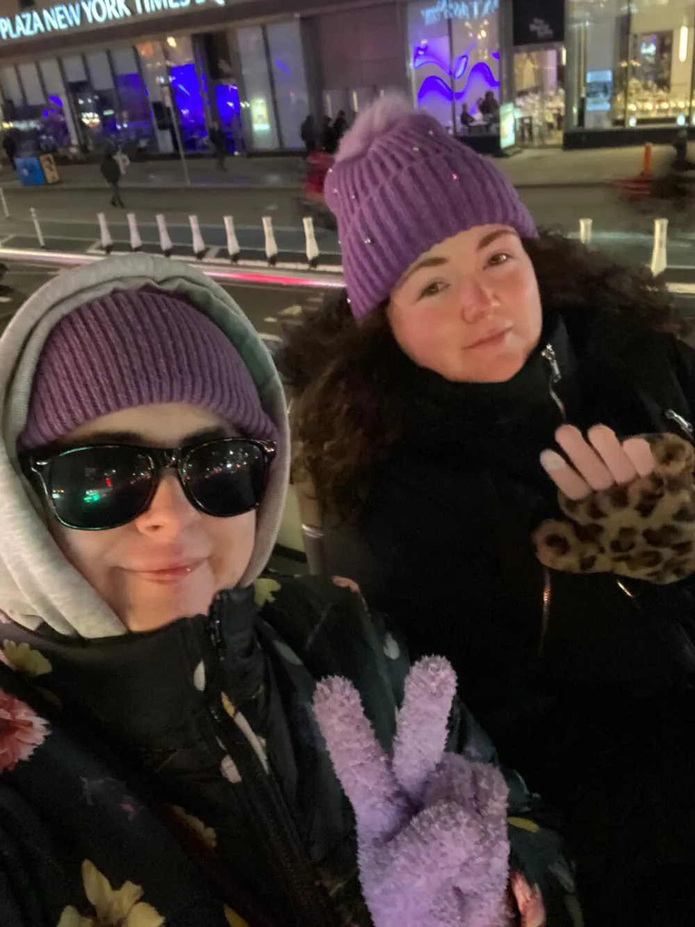 Two individuals are posing for a selfie on a city street at night wearing warm clothing and smiling with one making a peace sign gesture