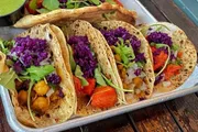 The image shows a tray of vibrant, vegetable-filled tacos with a bright purple cabbage garnish and a side of green sauce.